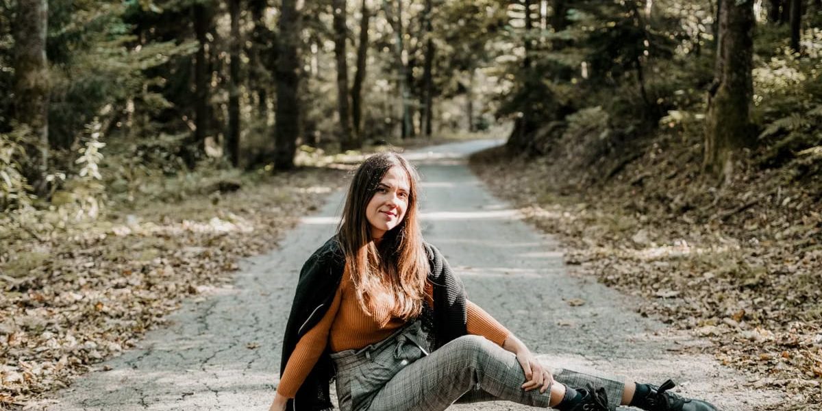 Cheerful young female tourist sitting on rough roadway between green trees in woods and looking at camera