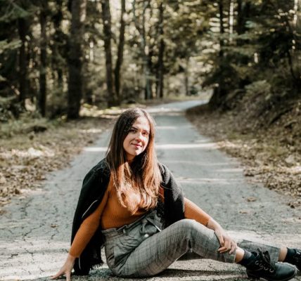 Cheerful young female tourist sitting on rough roadway between green trees in woods and looking at camera