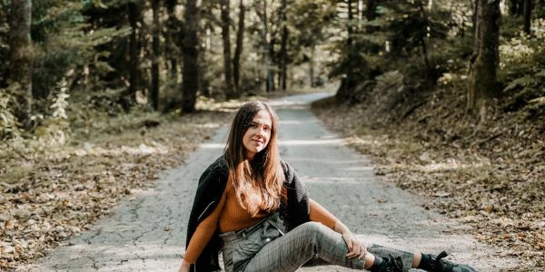 Cheerful young female tourist sitting on rough roadway between green trees in woods and looking at camera