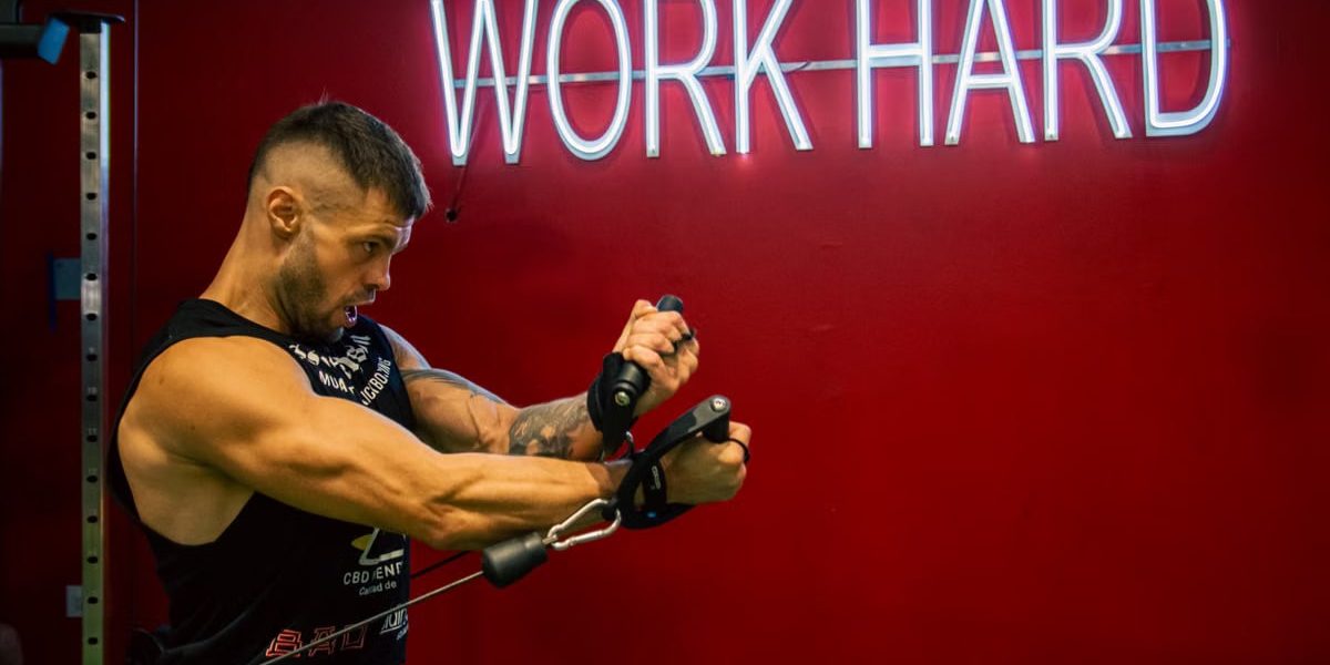 A muscular man exercising in the gym with a motivational 'Work Hard' neon sign on a red background.