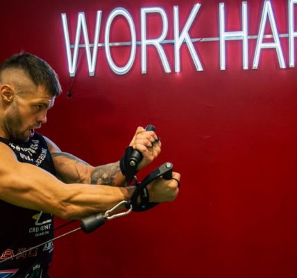 A muscular man exercising in the gym with a motivational 'Work Hard' neon sign on a red background.